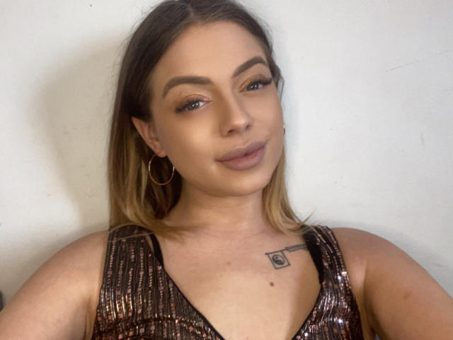 BritnyDesire with shoulder-length blonde hair, wearing a shiny brown dress, hoop earrings, and a small tattoo on her chest, smiling slightly. Her tits are partially visible.