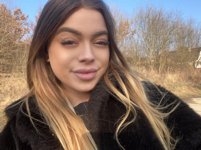 BritnyDesire with long brown and blonde ombre hair, black furry coat, and a slight smile, standing outdoors with dry grass and leafless trees in the background.