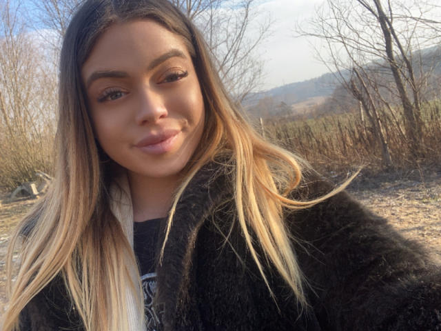 BritnyDesire with long blonde hair, brown eyes, and a subtle smile, wearing a black fur coat, standing outside in a sunlit, wooded area. Her tits are not visible.