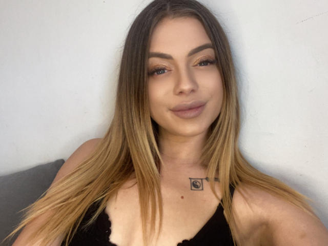 BritnyDesire with long blonde hair, black lace top, slight smile, tattoo on chest, brown eyebrows, subtle makeup, and soft pink lips. Boobs visible, arms relaxed.