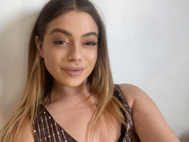 BritnyDesire has long blonde hair, brown eyebrows, and is wearing a sparkly brown dress, with hoop earrings, smiling softly at the camera. Her tits are partially visible.