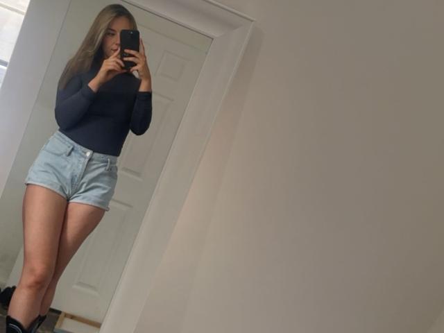 RoxiiGirl with long blonde hair, wearing a navy top, light blue shorts, and black boots, taking a mirror selfie, showing her ass and thighs.