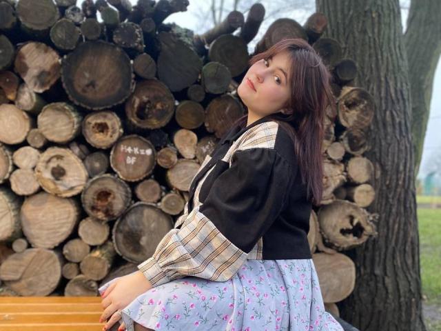 SpringChristie with brown hair, black and white striped shirt, and floral skirt, sitting on wooden bench, leaning back, showing her ass, in front of stacked firewood.