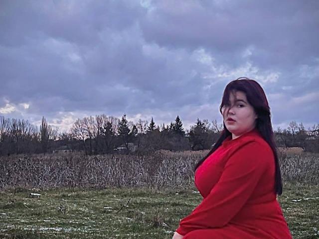 SpringChristie has large tits, wearing a red sweater, standing in a grassy field with a cloudy sky, dark hair, and a slightly confused expression.