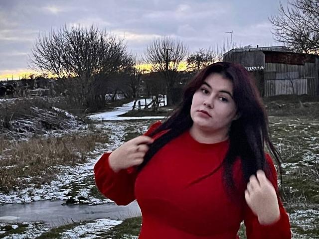 SpringChristie with long dark hair, red long-sleeve top, standing in a snowy, grassy area, hands near shoulders, looking slightly to the side, cloudy sky, bare trees in background.