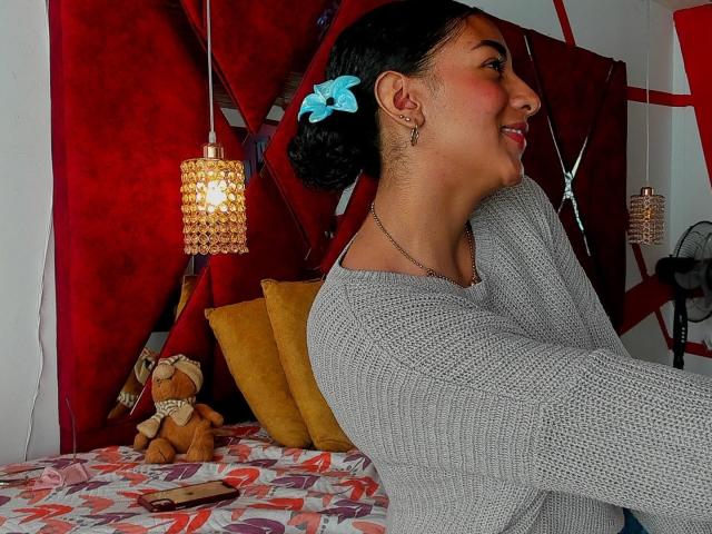 NatalyBlonder with dark hair in a blue bow, wearing a gray knit top, smiling sideways, showing her ass slightly, in a bedroom with red and yellow decor.