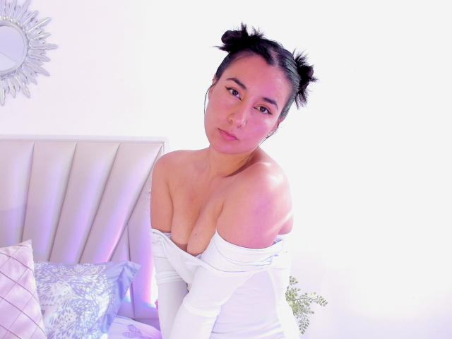 Nati69 avec des couettes, du fard à paupières rose, un haut blanc à une épaule, laissant entrevoir ses seins, assise sur un lit avec des coussins roses et blancs, regardant l'appareil photo..