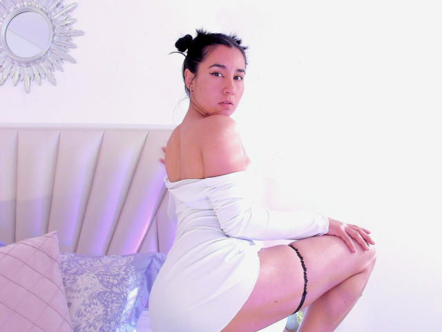 Natii69 trägt ein weißes Kleid mit offener Schulter, sitzt auf einem Bett, zeigt ihr Gesäß und einen schwarzen String, hat dunkles Haar zu einem Dutt gebunden und blickt mit ernstem Gesichtsausdruck nach hinten..