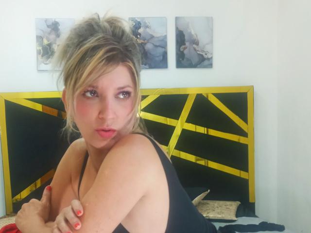 CoupleMature with blonde hair, pouty pink lips, and red nails, in a black tank top, sitting on a bed with yellow and black headboard, looking to the side.