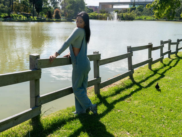 NicoColeman stands by a wooden fence, wearing gray overalls, exposing her ass, with long black hair, near a grassy park and a water-filled area with a fountain.