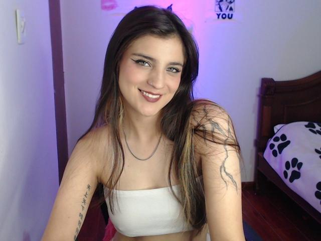 SarahhTanyas with long brown hair, smiling, wearing a white strapless top, showing a tattoo on her right arm, and a silver necklace, in a bedroom with a wooden bed.