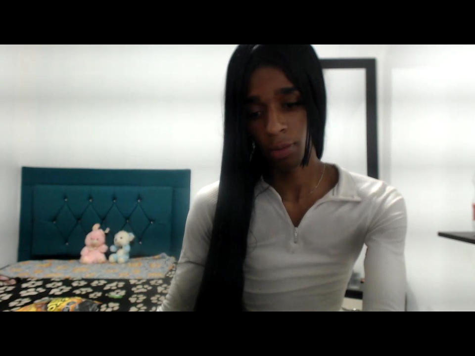 LucianaHottieX with long black hair, wearing a white polo, sitting, looking down, with a teal tufted headboard and stuffed toys in the background.