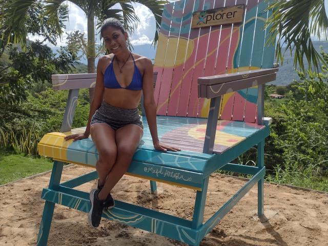 AllisonPearson with dark skin, blue bikini top, gray shorts, black shoes, sitting on colorful beach chair, smiling, tan, toned, small tits, black hair, tropical setting, sandy ground.