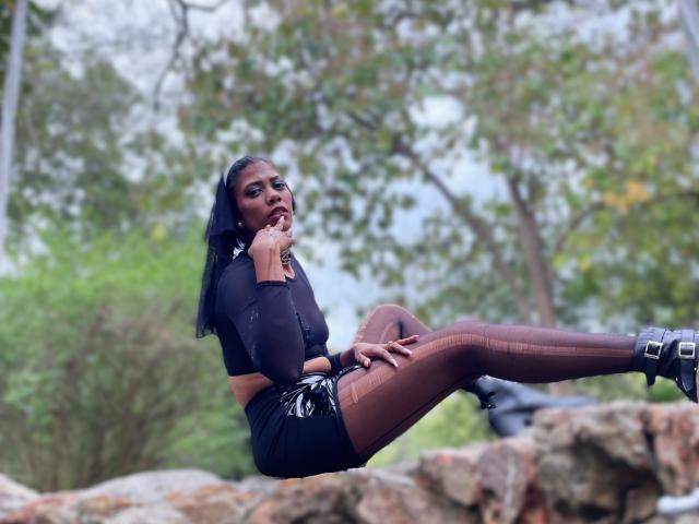 AllisonPearson with dark skin, black hair, and red lipstick, sitting on rocks, wearing black mesh top, shiny black shorts, sheer brown tights, and black boots, sucking her finger.