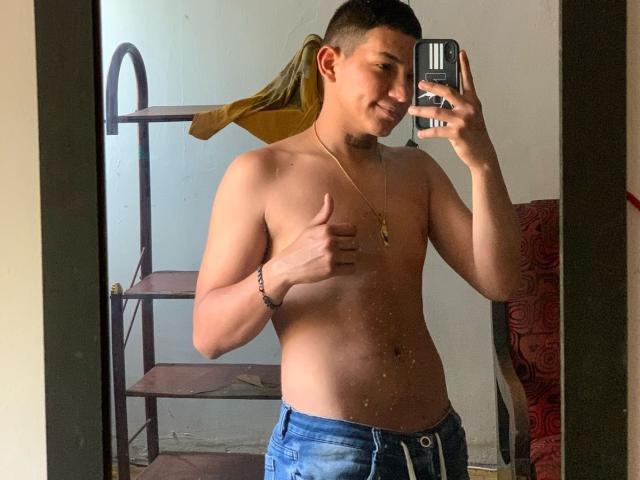 Shirtless DorianSexy with a ponytail, blue jeans, and a gold necklace, giving a thumbs-up in a mirror selfie, holding a phone. His ass is slightly visible.