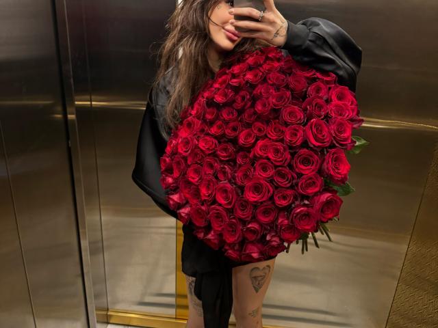 SensualMila with a huge bouquet of red roses, wearing a black oversized shirt, holding the roses against her chest, tattoos on her thigh, taking a mirror selfie.