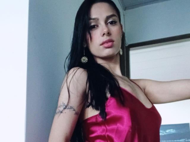 AnaisHilton with long black hair, pink satin dress, small tits, tattoo on left arm, gold earrings, serious expression, standing indoors.