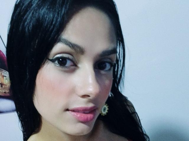 AnaisHilton has dark wet hair, brown eyes, thick black eyeliner, pink glossy lips, and a small nose with light skin. She wears yellow earrings and stares forward.