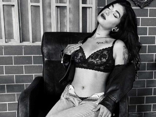NaomiDavis with dark lipstick, eyes closed, leaning back in a chair, wearing a lace bra, unbuttoned jeans, and a leather jacket, showing tattoos and a tattooed neck.