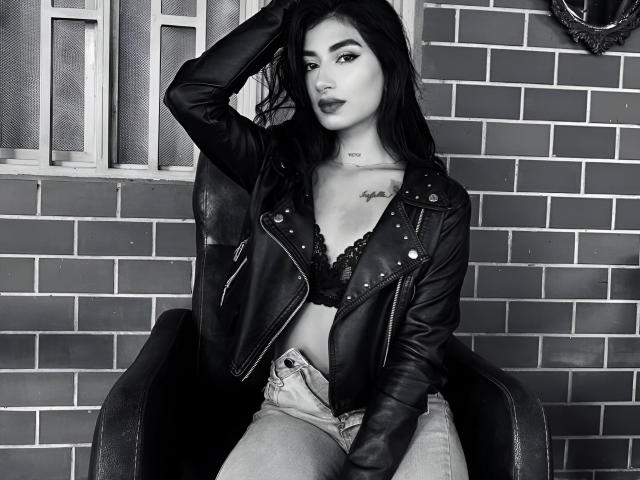 NaomiDavis with dark hair, wearing a black leather jacket, lace bra, and high-waisted jeans, sitting in a chair, hand in hair, tattoo on chest, sexy, black-and-white photo.