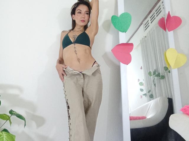Veroo69 with small tits, wearing a green bra and loose beige pants, standing in a room with colorful heart decorations, arm raised, looking at the camera.