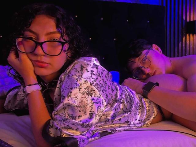VanillaHotG with curly black hair and glasses, wearing a floral dress, rests her head on her hand, next to a shirtless VanillaHotG with glasses and a watch.