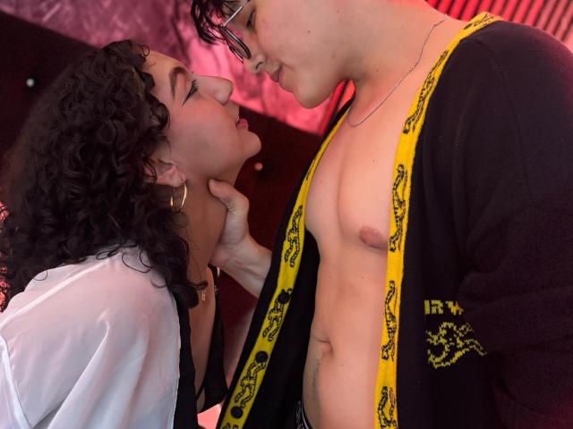 VanillaHotG with curly black hair and hoop earrings, holding the chin of a shirtless VanillaHotG with glasses, yellow and black robe, and visible nipples, in an intimate pose.