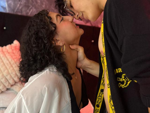 VanillaHotG with curly black hair, gold hoop earrings, white shirt, and black bra leans back as a guy with glasses and yellow tape over his shirt kisses her nose.