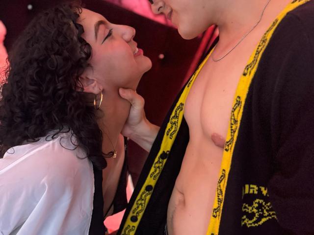 VanillaHotG with curly black hair, wearing a white shirt, leans in, eyes closed, as a shirtless VanillaHotG with a yellow and black robe holds her chin.