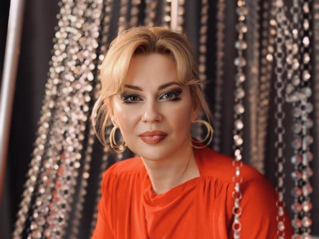 NatalyFoxy69 has blonde hair, green eyes, and wears a red dress with gold hoop earrings. She's looking at the camera with a subtle smile, showing her pouty lips.