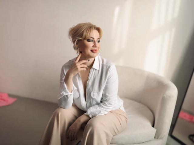 NatalyFoxy69, blonde, white shirt, beige pants, sitting on white chair, thinking with finger on chin, soft light, slight smile, pink item on floor, reflection on mirror.