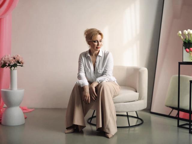 NatalyFoxy69 sits on a white chair, wearing a white shirt and beige pants, with hands on knees, blonde hair in an updo, looking to the side, in a sunlit room.
