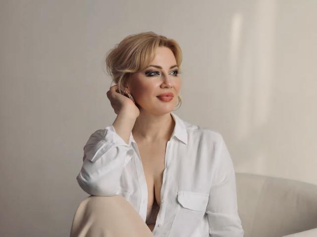 NatalyFoxy69 with blonde hair, wearing an open white shirt, showing her tits, sitting with one hand on her chin, smiling, in a soft-lit, minimalist setting.