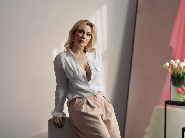 NatalyFoxy69 with blonde hair, white blouse, deep cleavage, and beige pants, leaning against a white surface, looking at the camera with a subtle smile. Her hands are in her pants.