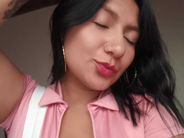 AradiaX with black hair, nose ring, and pink shirt, puckering lips, eyes closed, wearing gold hoop earrings, white backpack strap visible. Her ass is not in the frame.