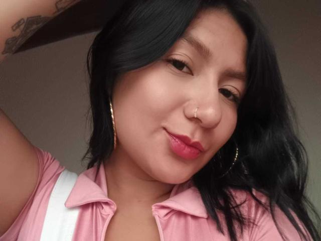 AradiaX has medium brown skin, black wavy hair, pink shirt, hoop earrings, nose ring, pink lipstick, slight smile, white strap, and a tattoo on her right arm.