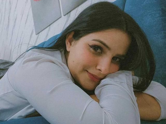 Ateneah, avec de longs cheveux noirs, une chemise blanche, allongée sur un canapé bleu, souriant à la caméra, les yeux légèrement plissés, les bras croisés sous le menton, un léger blush rose sur les joues, adorable..