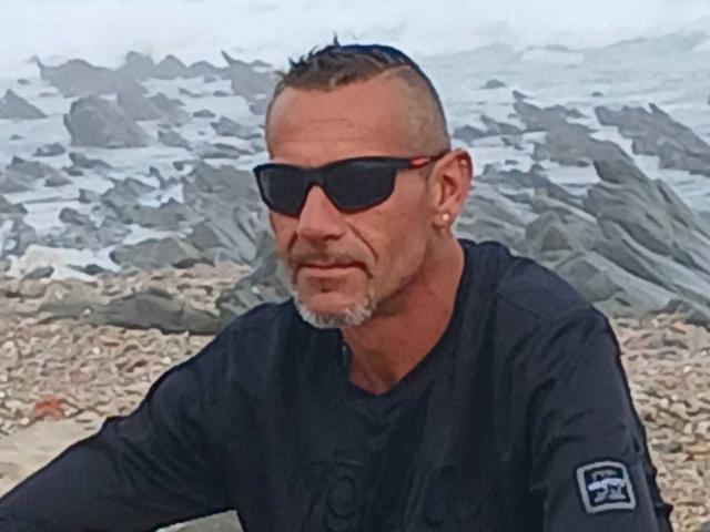 BobbyStar ha la testa rasata, occhiali da sole neri, una barba grigia e indossa una maglietta nera con una toppa. È seduto su una spiaggia rocciosa..