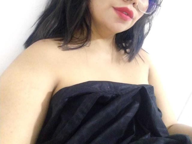 LilyRousee with black strapless dress, red lipstick, and glasses, showing bare shoulders and upper chest, with medium tits, fair skin, and black hair in a slightly messy style.