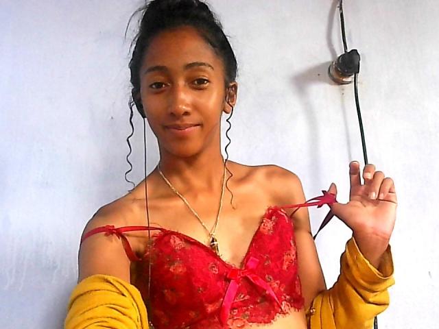 DiamondI with dark curly hair, wearing a red lace bra, yellow shirt, and gold necklace, holding a bra strap, smiling, showing small tits and a cute face.