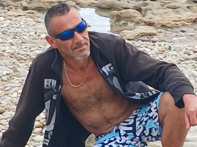 BobbyStar with gray hair and beard, wearing blue sunglasses, black jacket, and blue patterned swim trunks, has a hairy chest and a silver necklace.