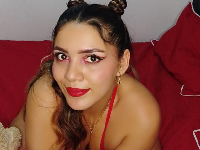 JuaniiSmiler has red lipstick, pink eyeshadow, and a double bun hairstyle. She's wearing a red halter top, sitting on red pillows with white cords. Her tits are partially visible.