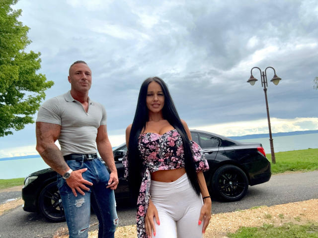 MyaAndDaniel with long black hair, floral off-shoulder top, white pants, standing next to a muscular man in a grey shirt and ripped jeans, in front of a black car.