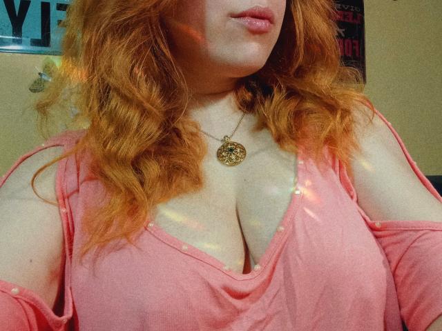 SpicyGrace with wavy red hair, pink off-shoulder top, and gold necklace, showing her large, shiny tits and pink lips in a slightly sunlit room.