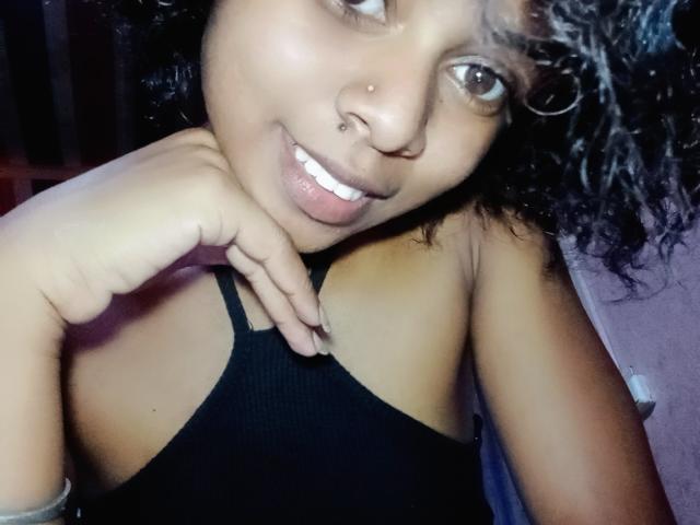 Zoriah has curly black hair, a nose piercing, and is smiling with her tongue slightly out. She's wearing a black tank top, showing her small tits.