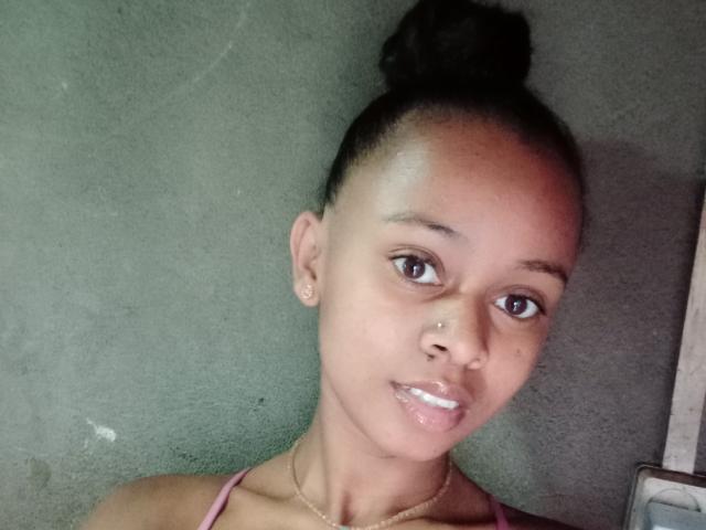 Zoriah has a dark brown bun, small nose piercing, and shiny lips, wearing a pink strap, looking at the camera with brown eyes. Her ass is not visible.
