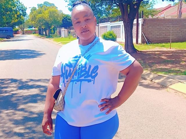 NteBobbyXx stands on a sunny street, wearing a white shirt with blue graffiti, blue pants, and a small crossbody bag, hands on hips, smiling confidently.