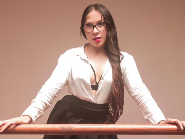 Kamie Carter, con cabello castaño largo, gafas, camisa blanca, sostén negro y falda negra, apoyada en una barra de madera, mostrando sus pechos y una expresión segura y sensual..