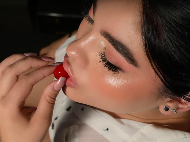AliceSamanta with shiny skin, black eyebrows, long eyelashes, pink lips, and black heart earrings, sucking on a red cherry, wearing a white shirt with black dots.