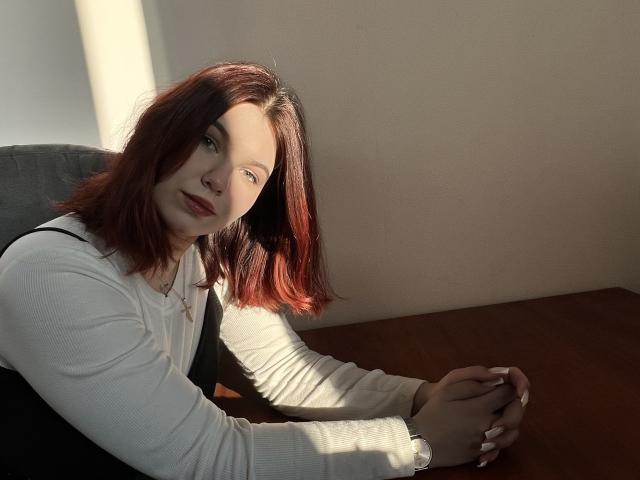 Khaleessi with red hair, white long-sleeve shirt, and black dress, sitting at a wooden table, sunlight on her face, hands clasped, subtle smile, nose ring, and long nails.